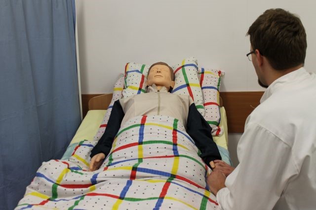 Opiekun medyczny siedzący przy łóżku, na którym leży manekin
