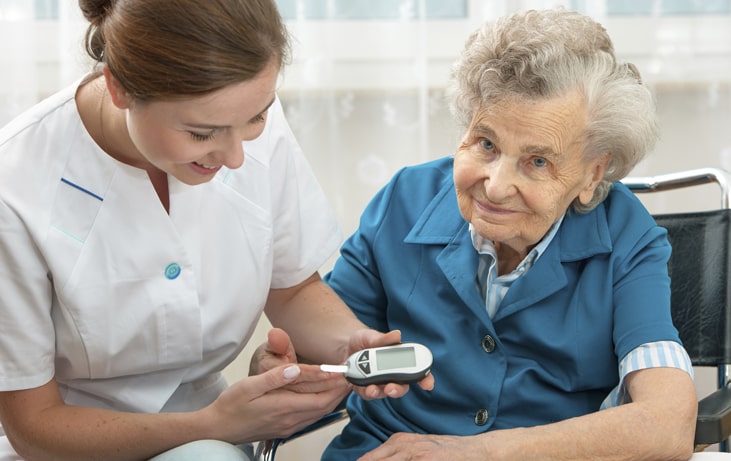 Opiekunka medyczna siedzi obok seniora i sprawdza jej poziom cukru przy pomocy urządzenia medycznego